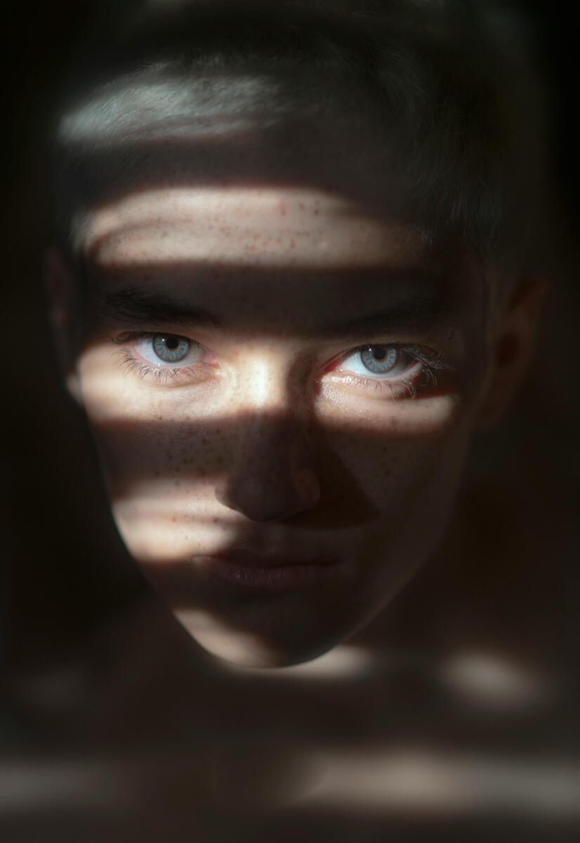 Gros plan sur le visage d'une personne aux yeux clairs et taches de rousseur, éclairée par des ombres portées de stores, photographie d'art minimaliste.